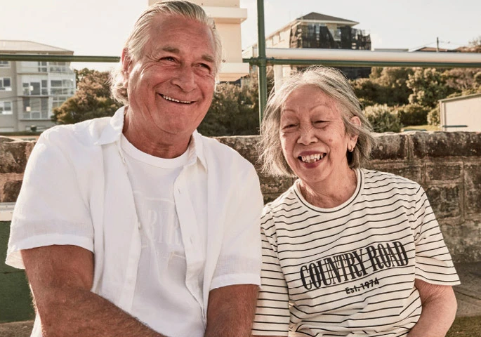 An older man in a white shirt and an older woman in a striped t-shirt smiling outdoors together for Country Road.