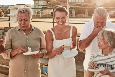 Group of four adults eating outdoors, holding small white plates and food in a sunlit setting