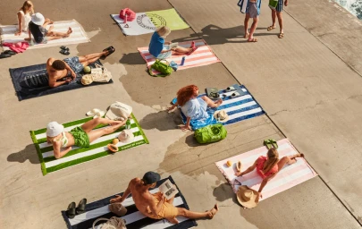High-angle aerial shot of several people relaxing on striped beach towels on concrete ground beside water.