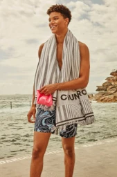 Smiling man standing near the ocean wearing patterned swim shorts and a striped towel draped over his shoulders