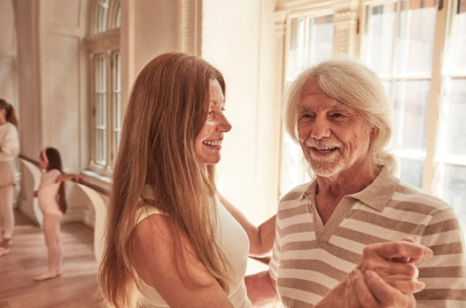 An older couple dancing together in a brightly lit room with arched windows.