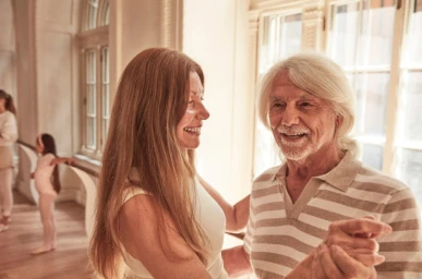 An older couple dancing together in a brightly lit room with arched windows.