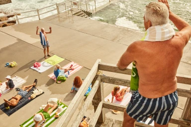 High-angle view of older man on wooden balcony overlooking people sunbathing and swimming near a concrete pool area