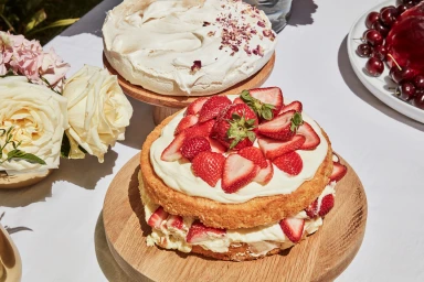Close-up overhead shot of a layered strawberry sponge cake on a wooden board with white roses and a pavlova in the background.