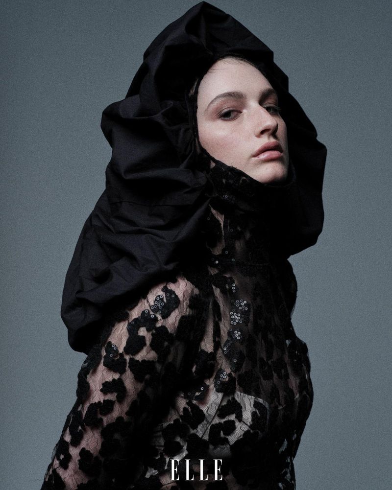 Portrait of a fair-skinned female model wearing a dramatic black hooded top with lace overlay in a studio fashion editorial shot.