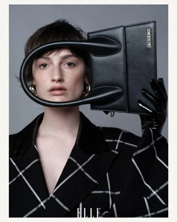 A woman in a black and white jacket poses with a black Jacquemus purse serving as a headpiece in a grey-toned photo studio.