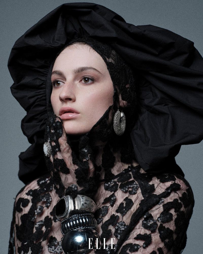 Portrait of a woman wearing a dramatic black lace and textile headpiece and matching top with chunky bracelets and hoop earrings.