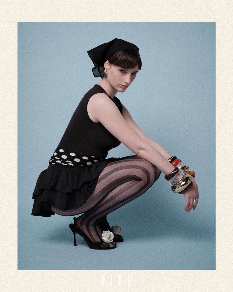 A young woman in a black top and ruffled skirt poses in a studio with a light-blue backdrop, wearing striped tights and high-heel shoes.