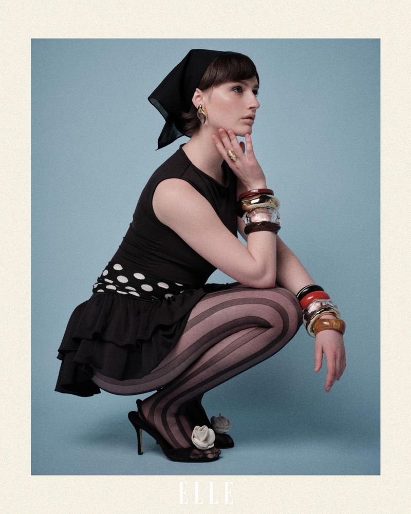 A woman in vintage-style black and white polka dot dress, scarf and high heels poses in a studio setting against a blue background.