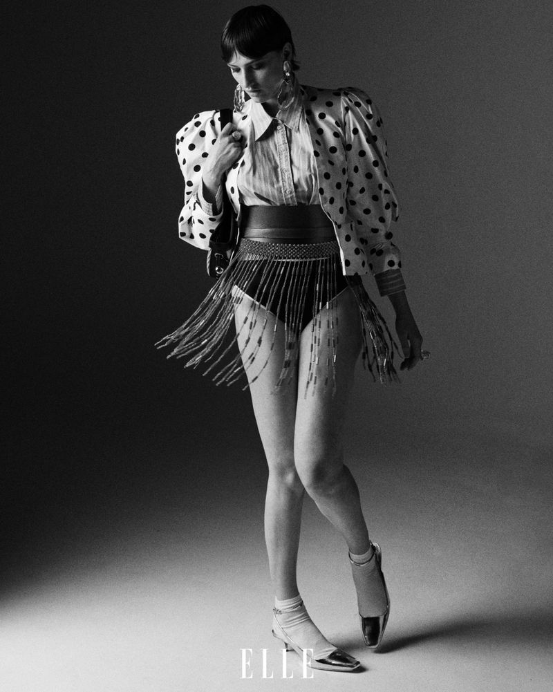 Full-length shot of a woman in a polka dot jacket, belt with fringe, and metallic shoes, posing in a grey studio.