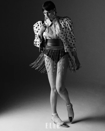 Full-length shot of a woman in a polka dot jacket, belt with fringe, and metallic shoes, posing in a grey studio.