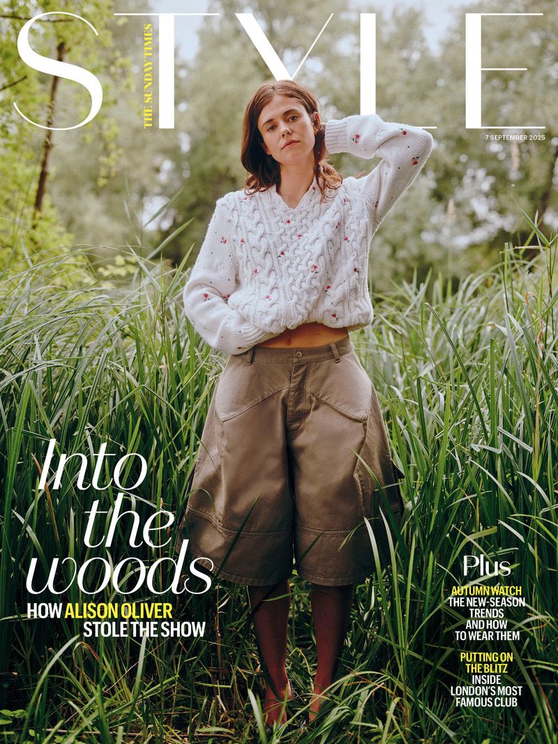 Magazine cover features a woman in a white sweater and brown culottes standing in a field of tall grass, with trees in the background.