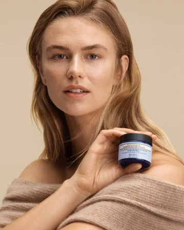 Woman in sweater holding jar of face cream in a studio shot for beauty product advertisement, showcasing skincare and complexion.