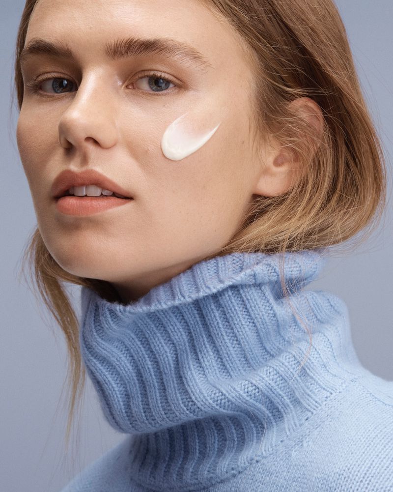 Close-up of a fair-skinned woman with blonde hair wearing a blue turtleneck, featuring a dab of white cream on her cheek.