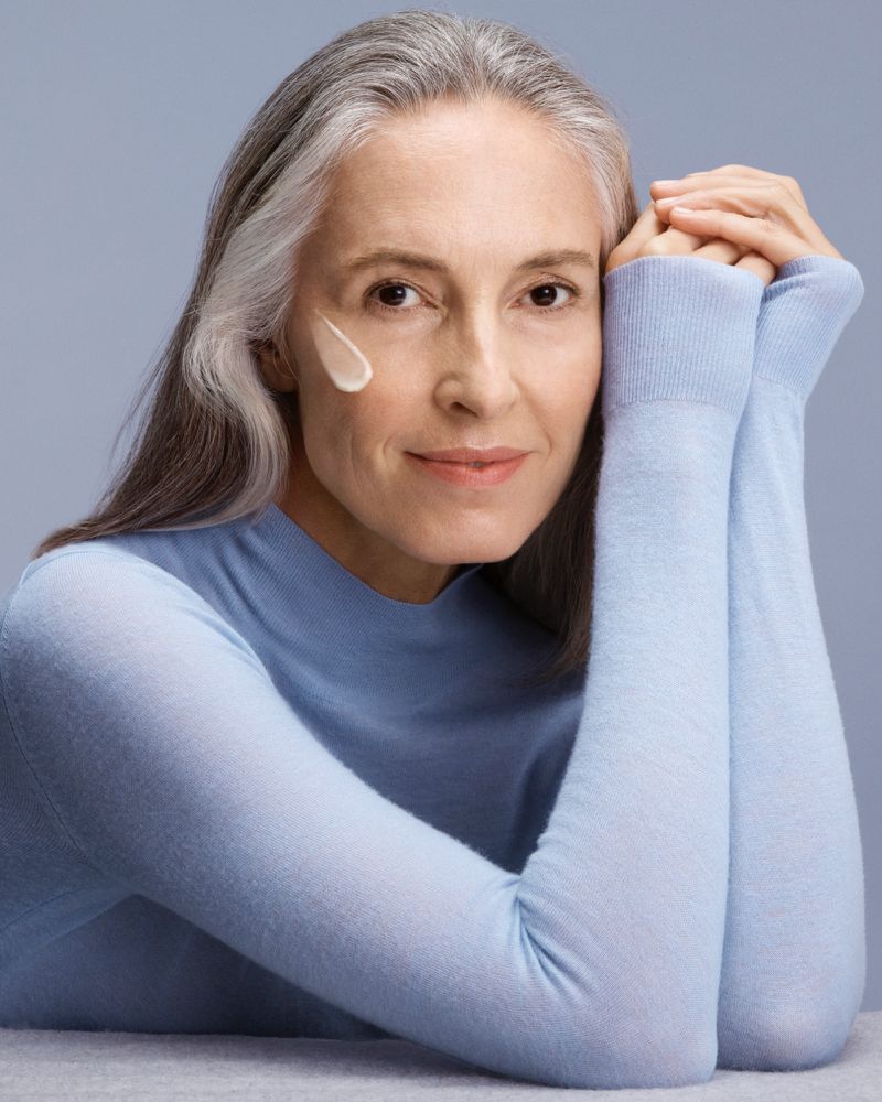 Mature woman with gray hair poses with hands near her face, showcasing a light moisturizer on her cheek in a beauty setting.
