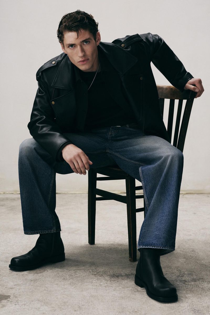 A young adult male model wearing a black leather jacket, black top, blue jeans, and black boots is seated in a neutral indoor setting.