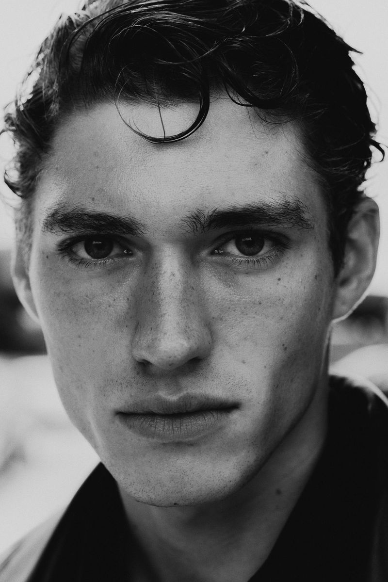 Black and white portrait of a young adult man with dark hair and eyes, shot in natural light with shallow depth of field.