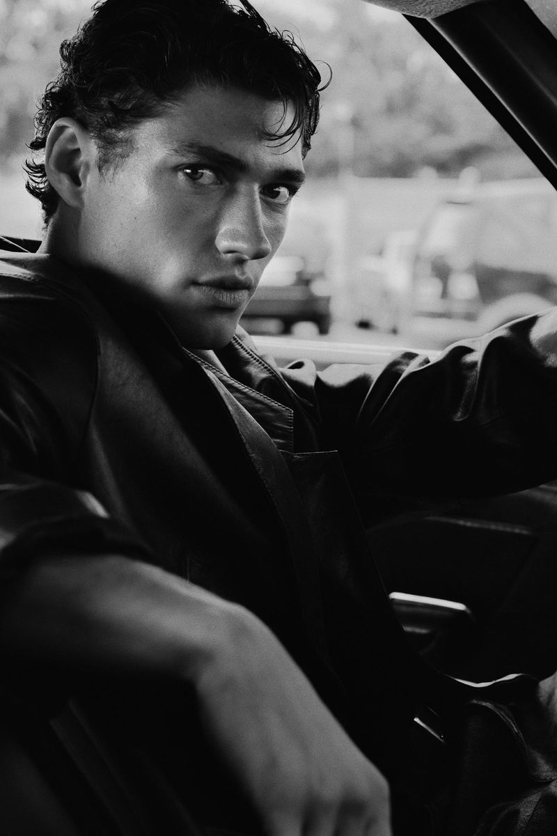Black and white portrait of a male model seated in a car, looking towards the camera, wearing a dark jacket and with an outdoor background.