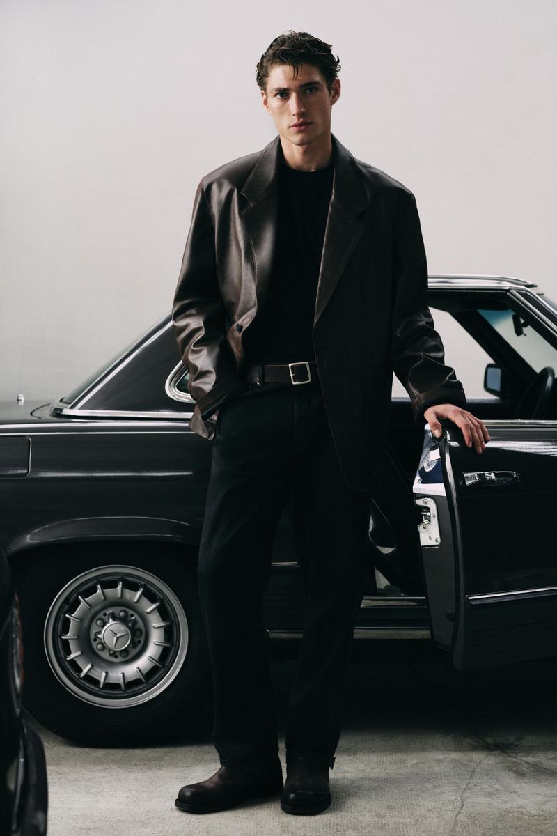 Man in black shirt and brown jacket posing next to black car with open door in studio. Fashion editorial style full shot.