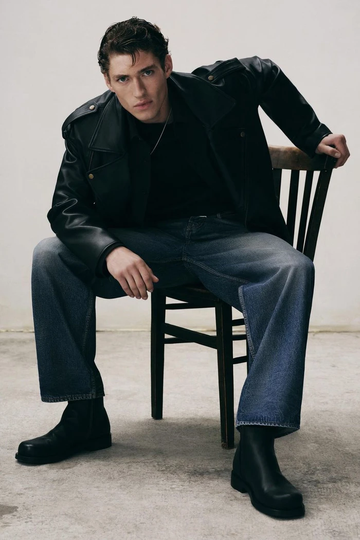 Model wearing a black leather jacket and blue jeans sitting on a wooden chair against a neutral background