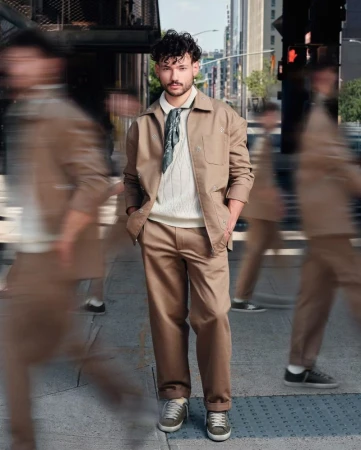 Man in beige jacket and trousers stands on city sidewalk amidst blurred pedestrians, with urban buildings in the background, fashion shot.