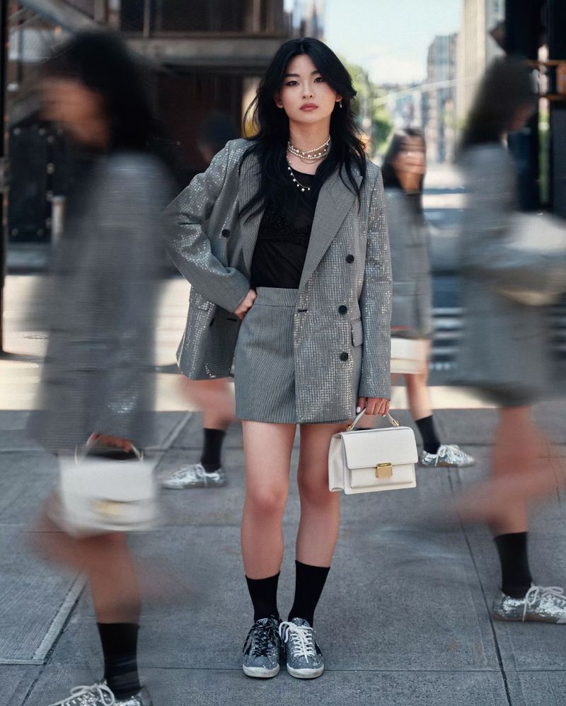 An Asian woman stands on a city street in a gray skirt suit, black top, and black socks, carrying a white purse with blurred pedestrians around her.