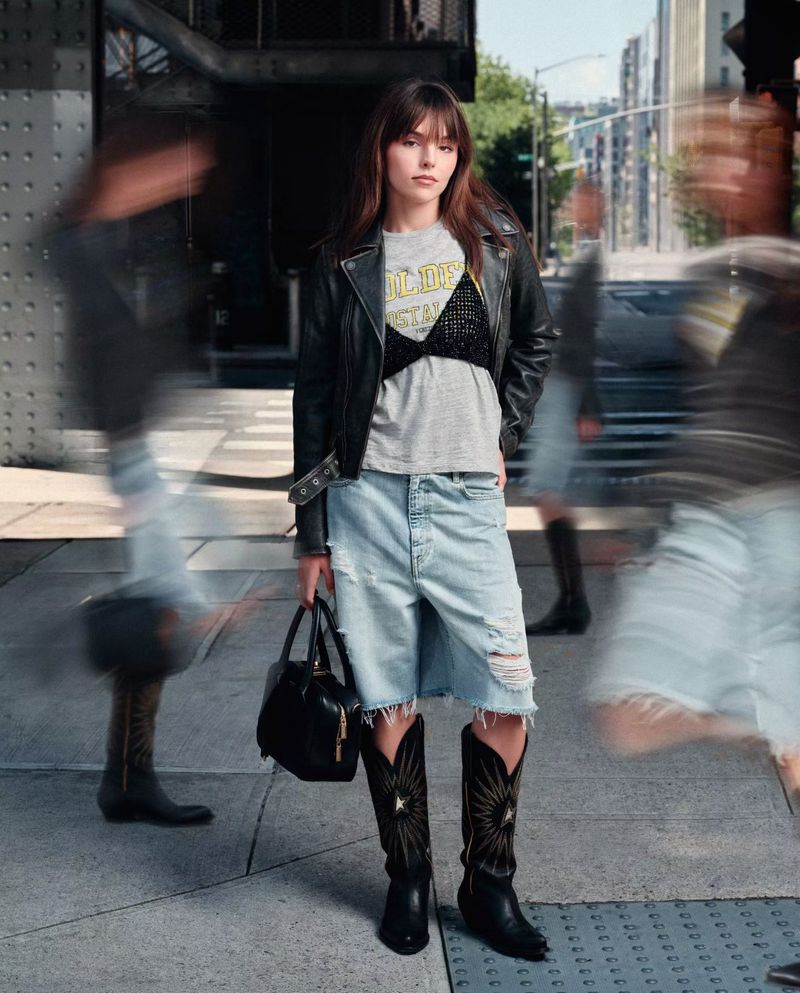 Fashionable woman in leather jacket, t-shirt, denim shorts, and cowboy boots poses on a city street with blurred people in the background.