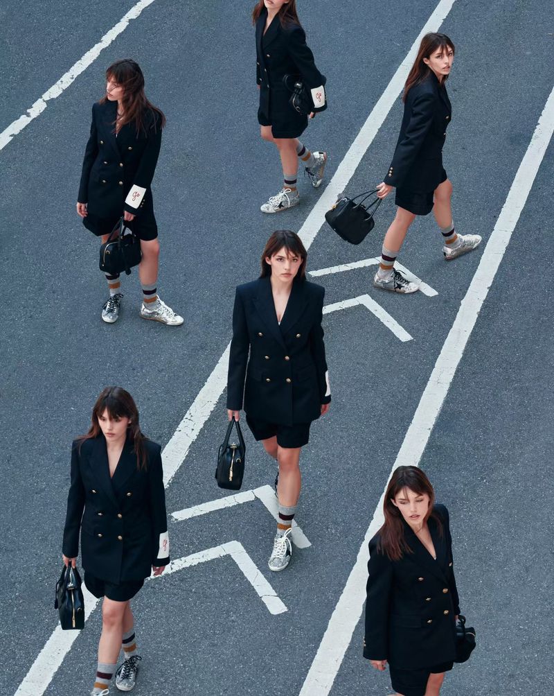 High-angle fashion shot of five adult women in black blazers and shorts, walking on an asphalt surface with white directional road markings.
