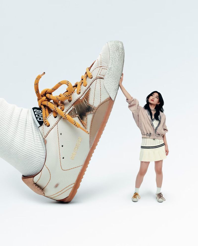 Oversized sneaker beside woman in tennis skirt and blush jacket, against a bright white background in a fashion shot.