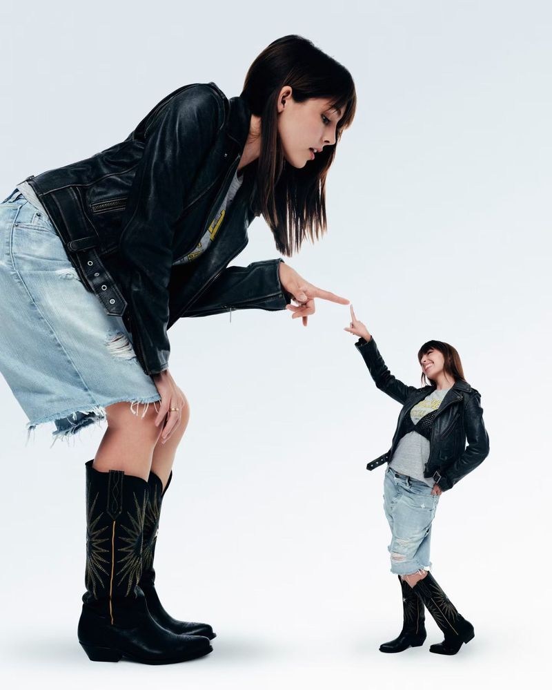 Two women pose in a photo studio against a white backdrop, wearing matching denim skirts, black leather jackets and cowboy boots.