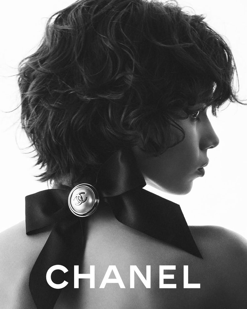 Monochrome fashion portrait of a young adult woman with wavy dark hair wearing a black bow on the back of her neck, logo visible.