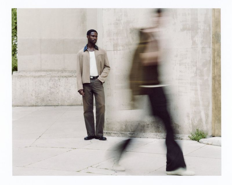 A Black man in a casual jacket and trousers stands outside a concrete building, alongside a blurred figure in motion, fashion editorial.