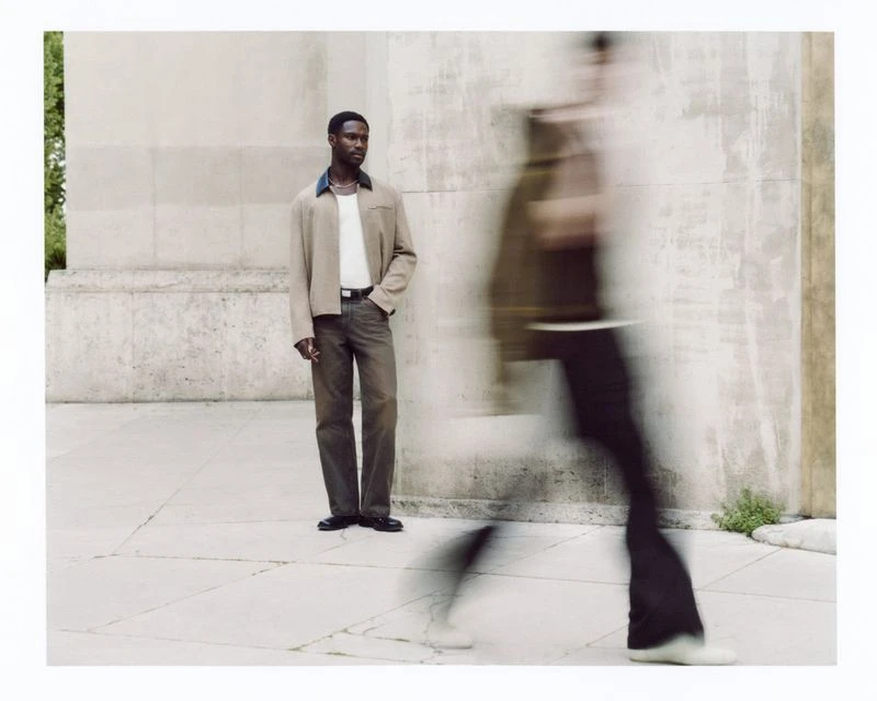 A Black man in a casual jacket and trousers stands outside a concrete building, alongside a blurred figure in motion, fashion editorial.