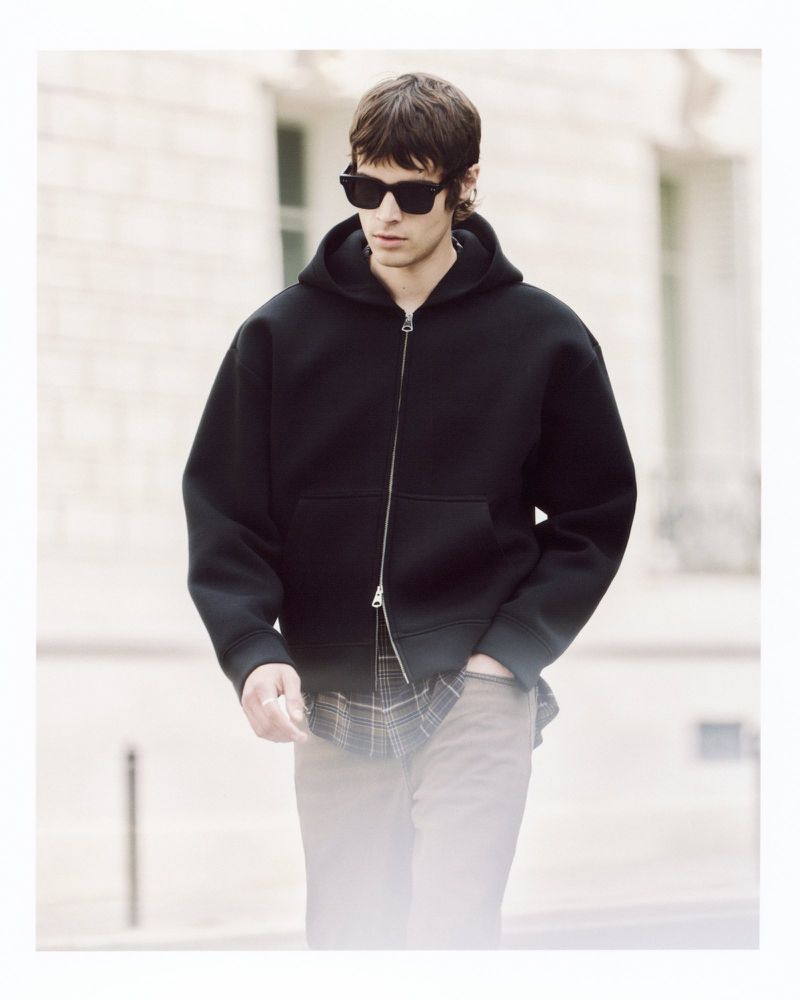 A young adult man walks on a city street wearing a black zip-up hooded sweatshirt, sunglasses, and brown pants in soft light.