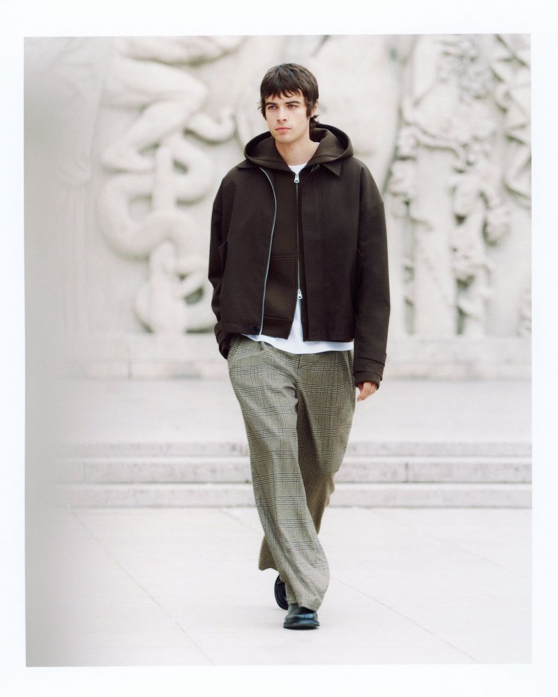 Male fashion model walks in a hooded jacket and loose plaid pants, posing in front of a classical stone wall with sculpture.