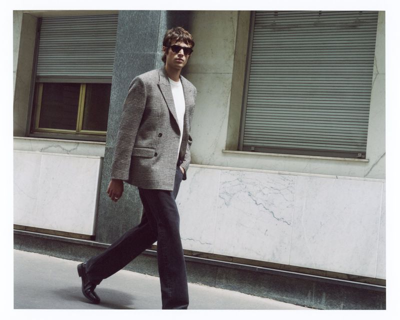 Model in sunglasses wearing a grey houndstooth double-breasted blazer and dark trousers walking along an urban sidewalk.