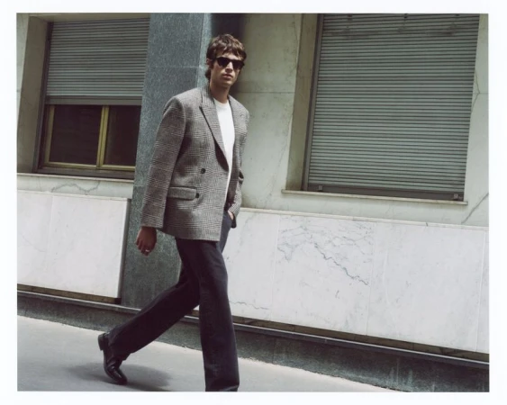 Model in sunglasses wearing a grey houndstooth double-breasted blazer and dark trousers walking along an urban sidewalk.