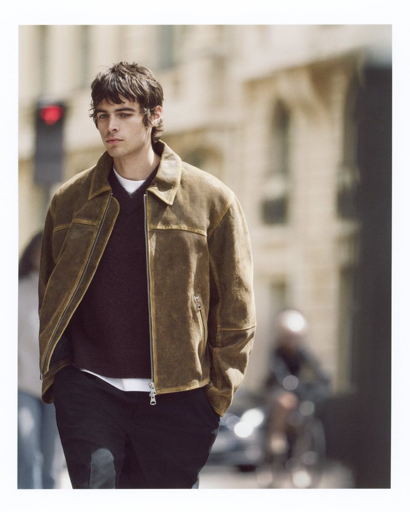 A young adult male in a brown suede jacket walks on a city sidewalk with blurred buildings and people in the background on a sunny day.