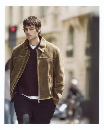 A young adult male in a brown suede jacket walks on a city sidewalk with blurred buildings and people in the background on a sunny day.