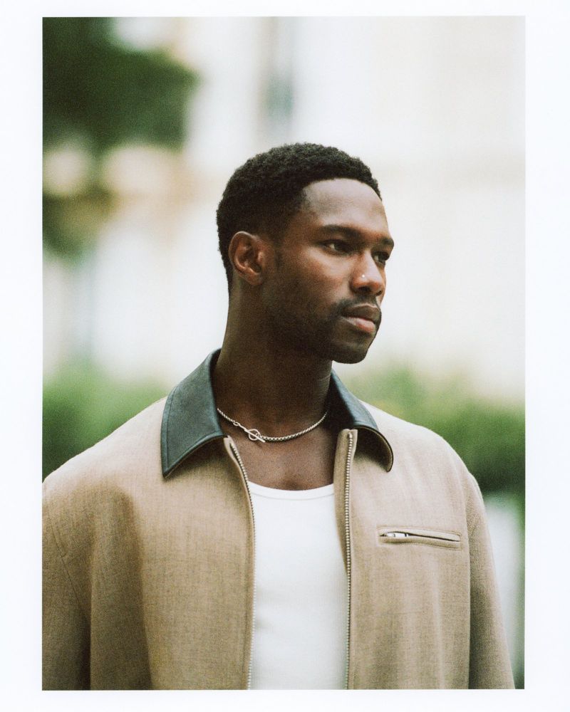 Portrait of handsome adult man with short dark hair in beige jacket and white t-shirt, shot outdoors in natural light against a blurred background.