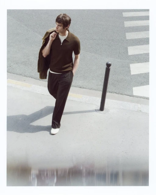 Man walking in brown polo and black pants with jacket over shoulder on crosswalk in sunlight for fashion or lifestyle editorial.
