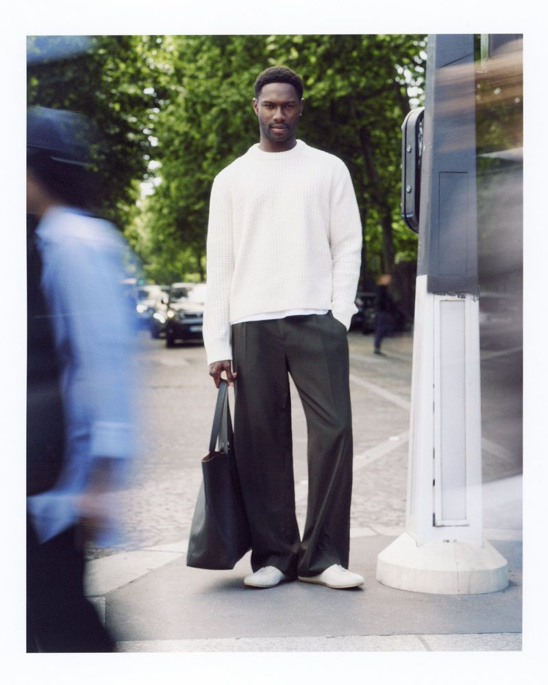 An African-American man in a white sweater and green pants stands on a city street holding a black tote bag in a lifestyle fashion shot.