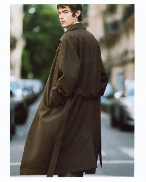 A young adult man in a long olive-green belted trench coat walking along a tree-lined city street with cars in the background.