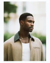 Portrait of a black man wearing a beige jacket with a dark collar and a silver chain necklace against a soft outdoor background.
