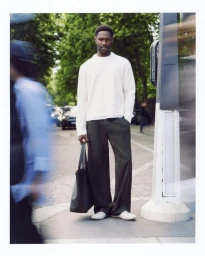 Model standing outdoors wearing a white knit sweater and dark trousers holding a large tote bag on an urban street.