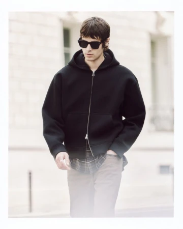 Model Tom Meacock wearing a black zip-up hoodie, dark sunglasses, and a layered plaid shirt while walking outdoors on a city street.