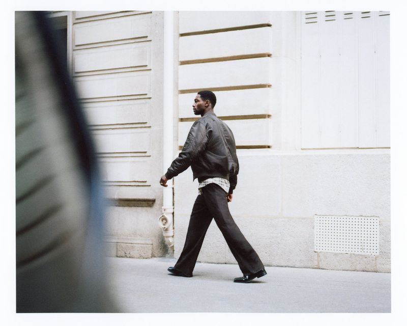 Model Sadiq Desh walks along an urban street wearing a dark bomber jacket and flared trousers for H&M Mens F/W 2025.
