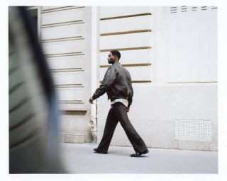 Model Sadiq Desh walks along an urban street wearing a dark bomber jacket and flared trousers for H&M Mens F/W 2025.