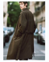 Model wearing an oversized dark brown trench coat looking over his shoulder on a city street.