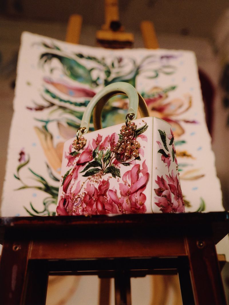 Floral-patterned white handbag with a jade handle displayed on a dark wooden easel in front of a painting in an indoor setting.